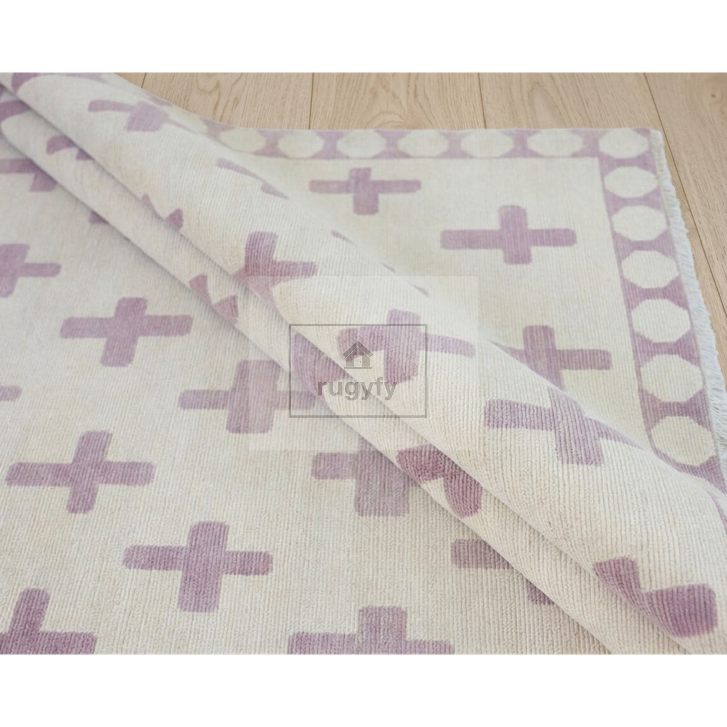 A cream-colored area rug with a lavender-purple geometric pattern of crosses and a matching dotted border, shown partially rolled over on a light wood floor. A watermark for 'rugty' is visible in the center.