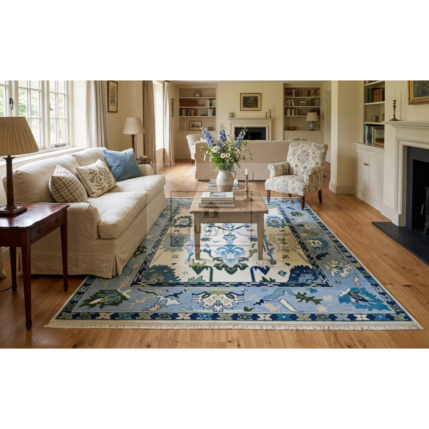 A large traditional-style area rug with a blue, beige, and white floral pattern is displayed in a bright, classic living room with hardwood floors, a beige sofa, and a wooden coffee table.