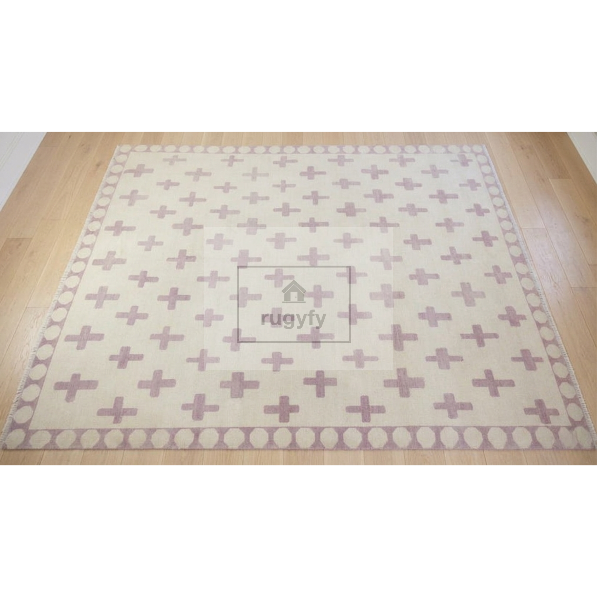 A rectangular area rug with a cream background and a repeating dusty pink cross pattern, bordered by pink and cream circles, shown on a light wood floor. A watermark for 'rugify' is visible in the center.