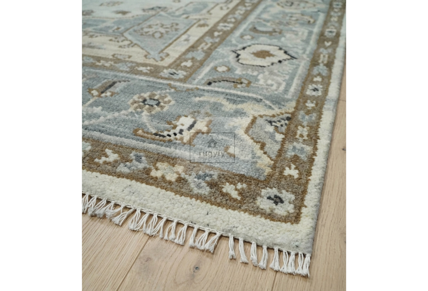 A close-up of an area rug with a traditional geometric pattern in muted blue-gray, beige, and brown tones, featuring a fringed edge on a light wood floor.