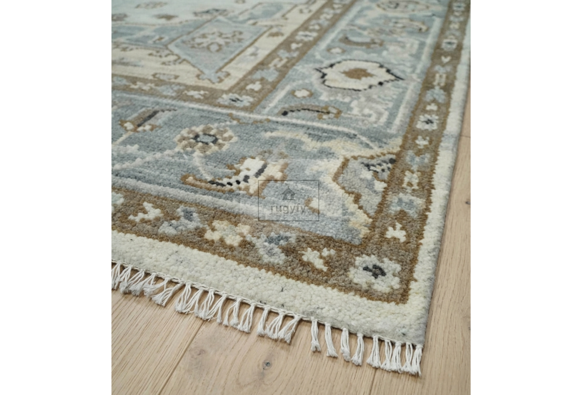 A close-up of an area rug with a traditional geometric pattern in muted blue-gray, beige, and brown tones, featuring a fringed edge on a light wood floor.