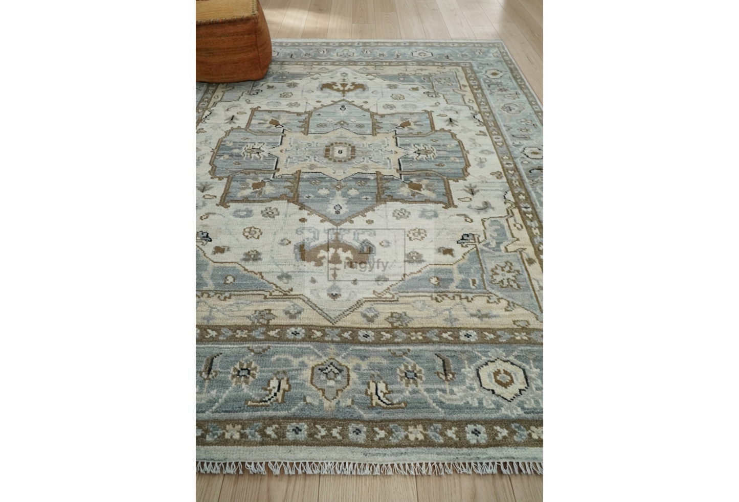 A rectangular area rug with a traditional medallion pattern in light blue, beige, and brown, shown on a light wood floor with a small portion of a wooden stool visible in the top left corner.